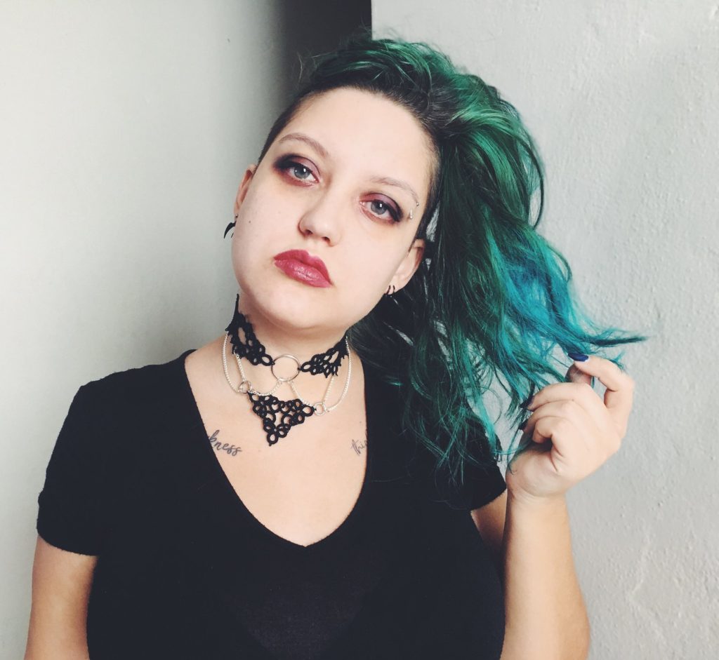 Image of Sparrow posing in front of the camera, chest up.  She is wearing a tatted lace collar that is connected by chains.  Her left hand touches som e of her blue-green hair.