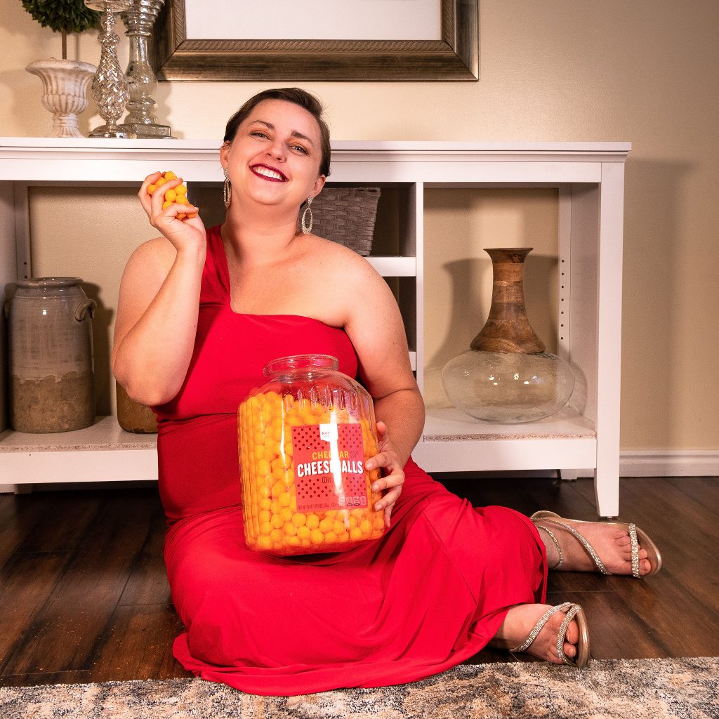 Plus size fashion model in red gown for in-home photoshoot, holding cheese puffs - we love snacks!