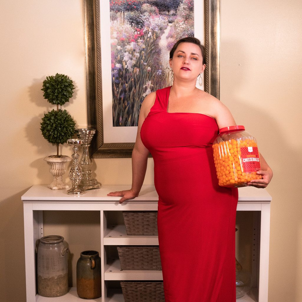 Plus size fashion model from Utah wearing a floor length red gown, holding cheese puffs as a snack for an in-home fashion editorial photoshoot in her living room