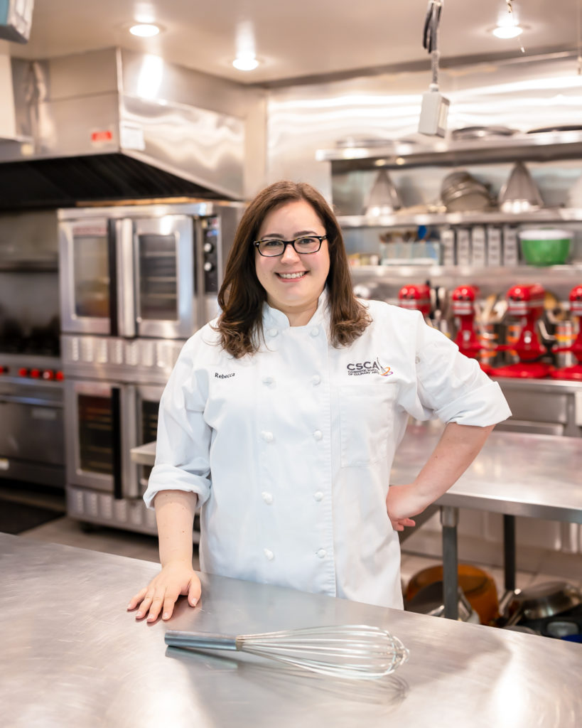 Rebecca is shown with her hair down, wearing glasses in a white chef coach.  She's in a professional kitchen with lots of gleaming steel appliances behind her.  

A large whisk is on the table in front of her.
