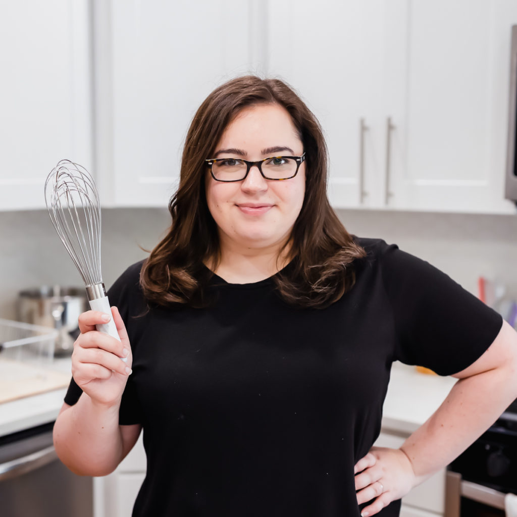 Rebecca is pictured in a ktichen, holding a whisk. Her brown hair is down, she's wearing glasses and has. a hand on her hip.  She's wearing a black t-shirt.