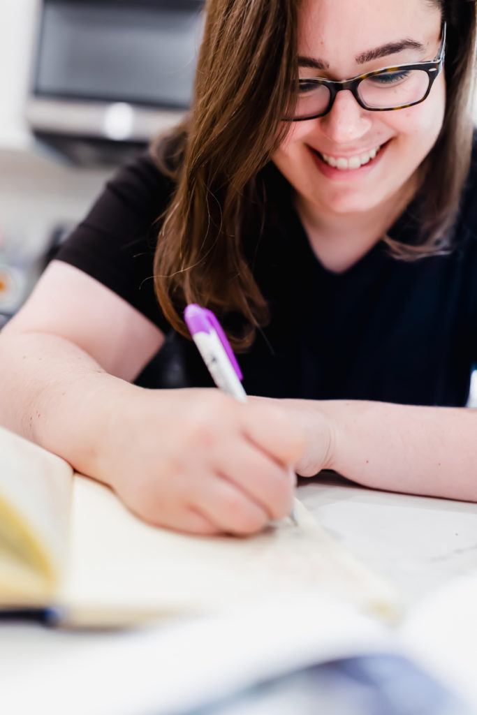 Rebecca is smiling as she writes in a notebook, planning recipe testing.  Her hair is down, she's wearing glasses and wearing a black-shirt.