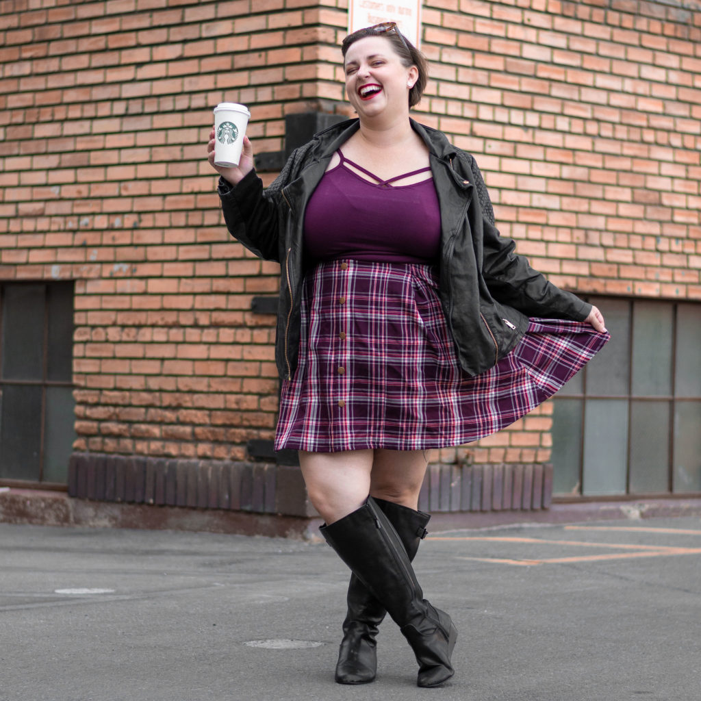 plus size blogger in purple plus size plaid skirt with knee high boots, a leather jacket, and purple cami.  She's holding a Starbucks cup in her hand