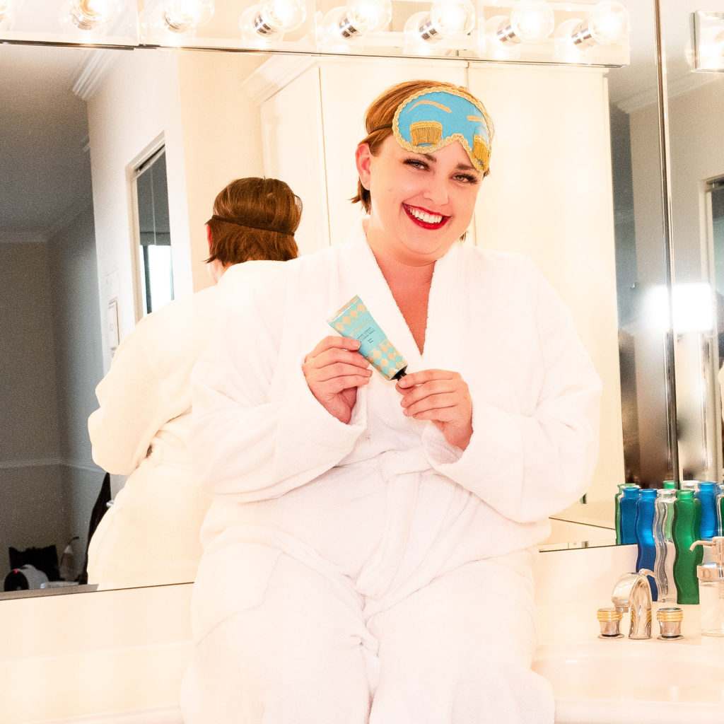 Brianne sitting on a bathroom counter in a white robe, holding a bottle of lotion with the iconic bright blue eye mask that Audrey Hepburn wore as Holly Golightly in Breakfast at Tiffany's!